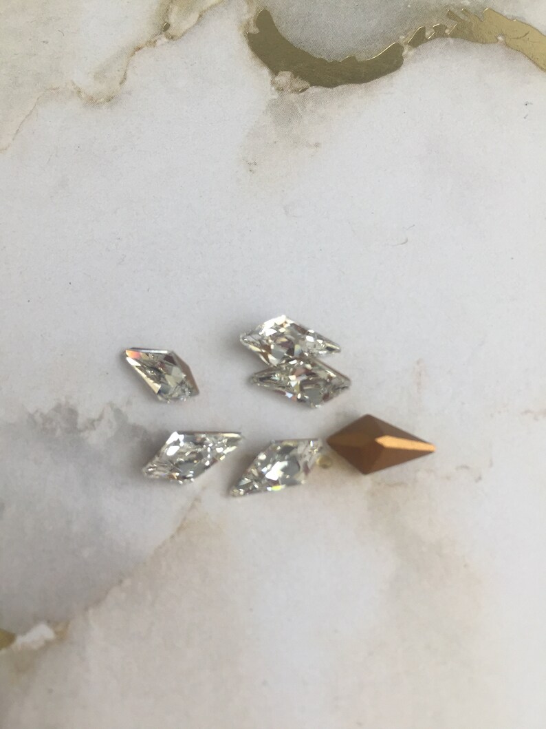 May include: Assortment of clear and gold rhinestones in various shapes, including a diamond shape, a teardrop shape, and a rectangular shape.
