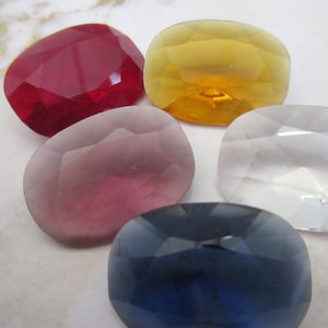 May include: Five faceted gemstones in red, yellow, lavender, clear, and dark blue. Each stone has a rounded rectangular shape with multiple facets, creating a sparkling effect. The gemstones are arranged closely together.