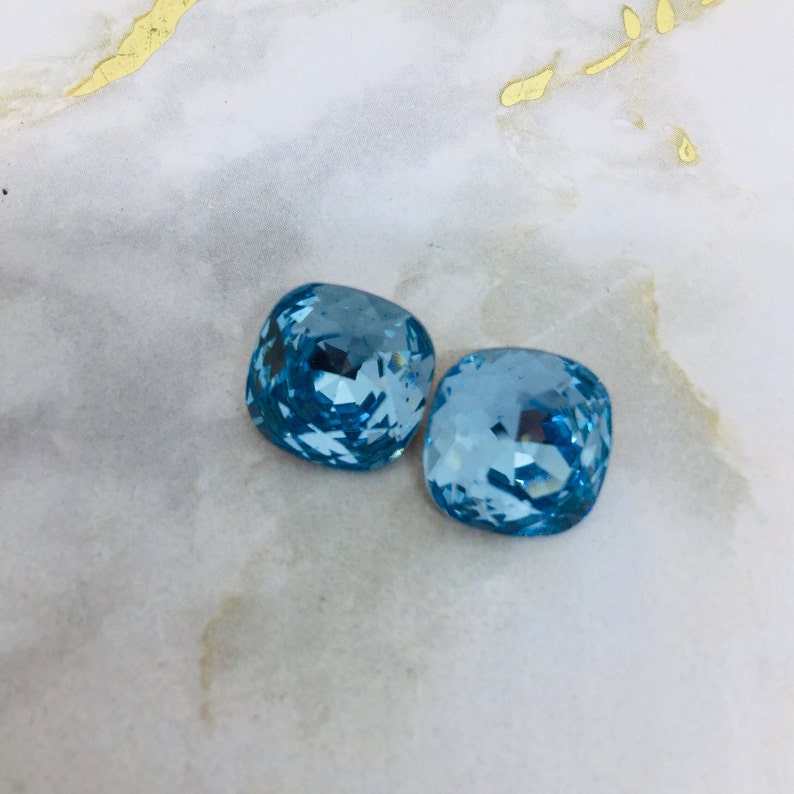 May include: Two light blue square-shaped crystal rhinestones.