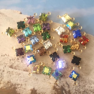 Könnte beinhalten: Eine Sammlung quadratisch geschliffener, mehrfarbiger Kristallanhänger mit goldfarbenen Metallschlaufen. Die Kristalle sind in verschiedenen Farben, darunter Grün, Blau, Lila und Rot. Die Anhänger sind auf einer hellen Oberfläche verstreut.