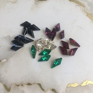 May include: Assortment of colorful rhinestones in various shapes and sizes. The rhinestones are arranged on a white marble surface. The rhinestones are in shades of green, blue, black, and purple.