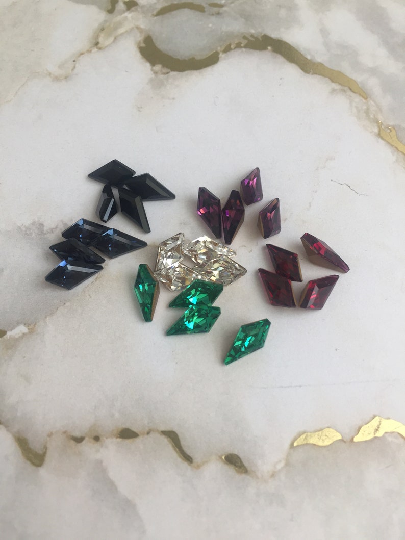 May include: Assortment of colorful rhinestones in various shapes and sizes. The rhinestones are arranged on a white marble surface. The rhinestones are in shades of green, blue, black, and purple.
