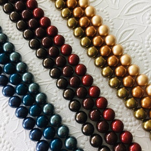 May include: Four strands of round, iridescent pearls in shades of teal, burgundy, brown, and gold. The pearls are arranged in rows on a white background.