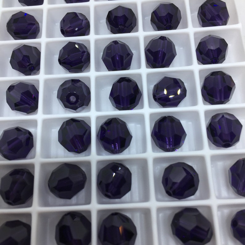 May include: A white plastic tray filled with 25 dark purple glass beads. The beads are arranged in a grid pattern.