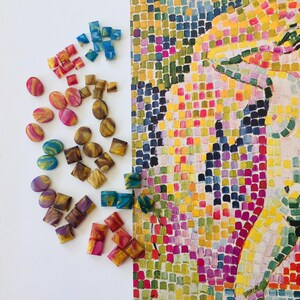 May include: An overhead shot shows a collection of colorful, handcrafted resin beads and mosaic art. The beads are in various shapes, including ovals and squares, and feature swirls of color. The mosaic art is a portrait with a vibrant, pixelated design.