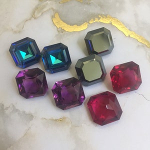 May include: Assortment of square-shaped gemstones in various colors, including purple, red, blue, and black. The gemstones are displayed on a white marble surface.