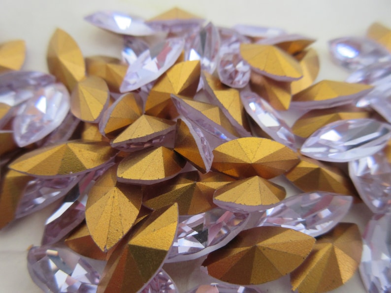 Puede incluir: Un mont&oacute;n de piedras de cristal transparentes, facetadas, ovaladas e iridiscentes con un revestimiento met&aacute;lico dorado en la parte posterior. Las piedras de cristal est&aacute;n esparcidas sobre una superficie blanca.