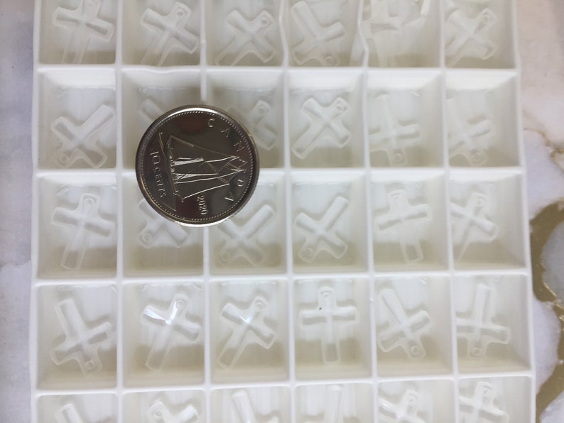 May include: A white plastic mold with 25 individual compartments, each containing a raised cross shape. A Canadian 10-cent coin is placed in the center of the mold.