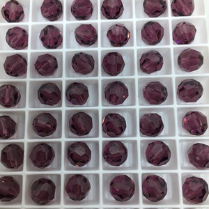 May include: A close-up view of a tray of round, faceted, purple glass beads. The beads are arranged in a grid pattern in a white plastic tray.