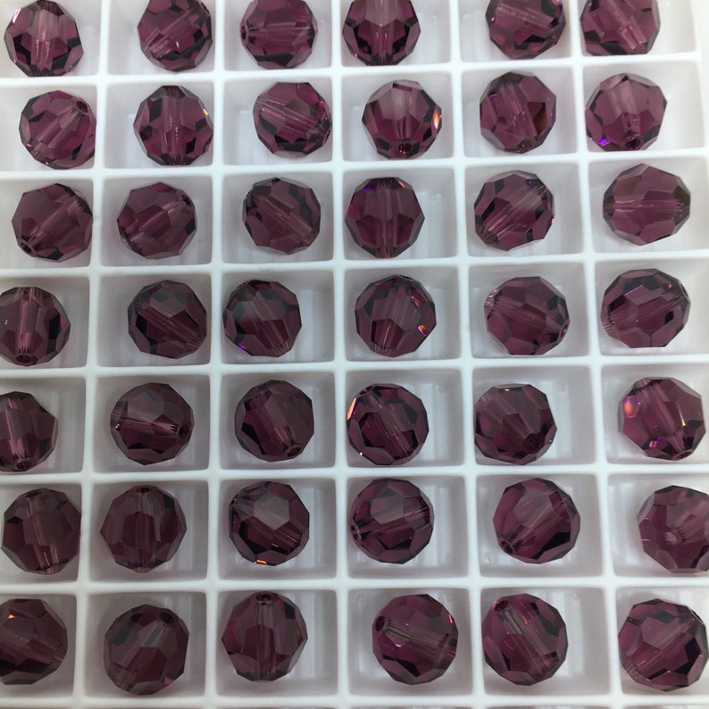 May include: A close-up view of a tray of round, faceted, purple glass beads. The beads are arranged in a grid pattern in a white plastic tray.