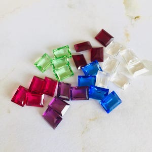 May include: Assortment of colourful rectangular glass gems in shades of green, red, blue, purple, and clear. The gems are scattered on a white surface.