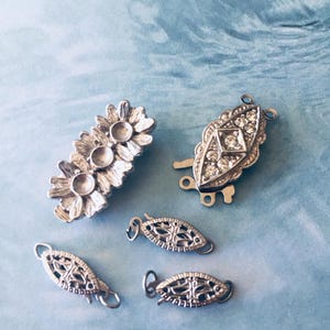Puede incluir: Un surtido de cierres y conectores de joyería plateados. Los cierres presentan varios diseños, incluyendo elementos florales, de filigrana y con pedrería. Las piezas están dispuestas sobre una superficie azul claro, mostrando sus intrincados detalles.
