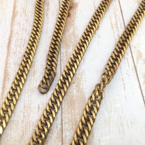 May include: Close-up of several gold-colored curb chains. The chains are arranged diagonally across a light-colored, wood-grain surface. The links are thick and closely interlocked, creating a textured appearance. The chains appear to be made of metal.
