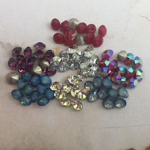 Könnte beinhalten: Sortiment aus bunten Glas-Straßsteinen in verschiedenen Formen und Größen. Die Strasssteine sind in einem Kreis auf einer weißen Oberfläche angeordnet. Die Farben umfassen Rot, Blau, Grün, Lila und Silber.
