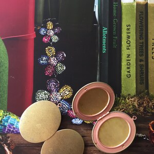 May include: Two round gold-coloured metal locket-style pendants with a third open locket pendant with a pink rim. The lockets are on a wooden surface with a book in the background.