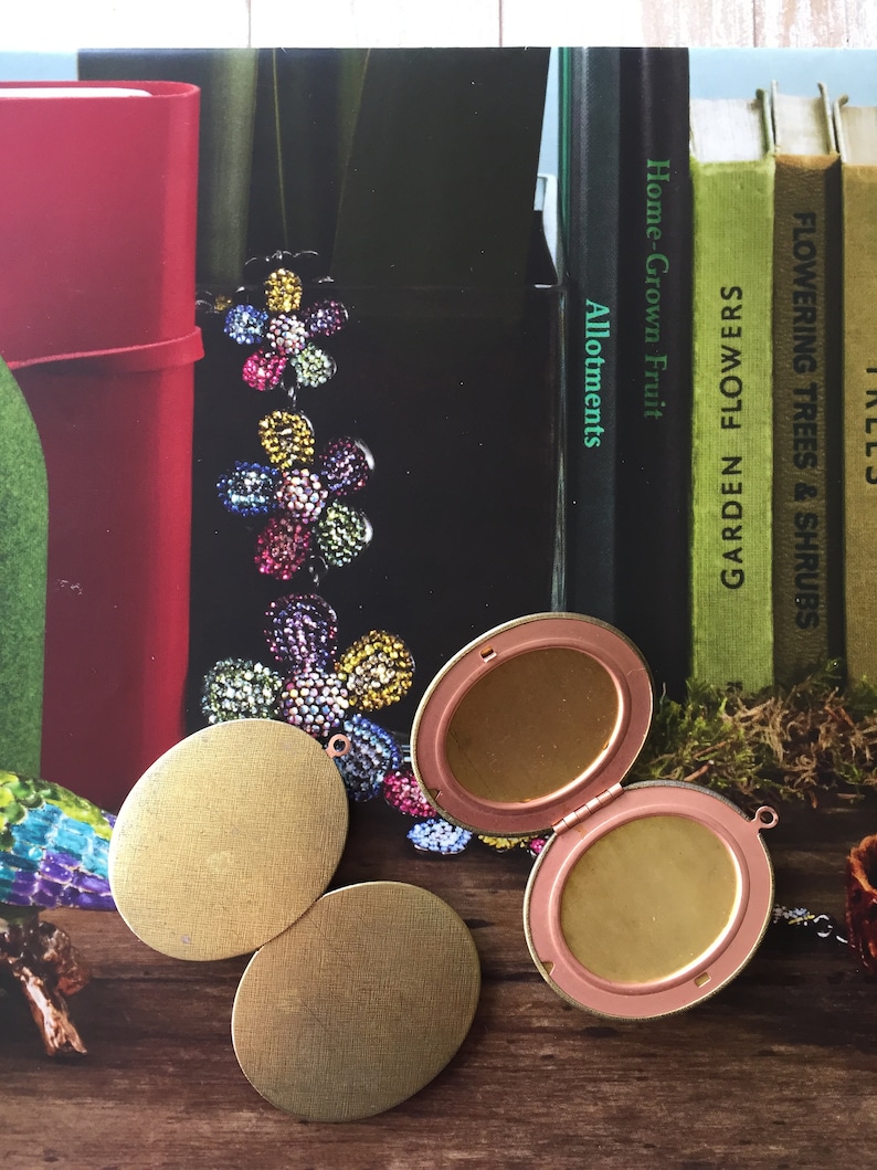 May include: Two round gold-coloured metal locket-style pendants with a third open locket pendant with a pink rim. The lockets are on a wooden surface with a book in the background.