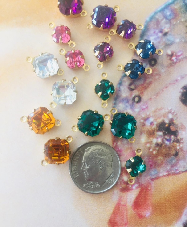 May include: A collection of colorful rhinestone connectors in various shapes and sizes. The gemstones are in shades of purple, pink, blue, green, orange, and clear. Each stone is set in a gold-tone metal setting with loops for attaching to other components.