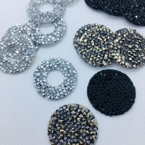 Puede incluir: Un surtido de adornos decorativos brillantes. La imagen presenta círculos y formas redondas cubiertas de pedrería plateada, negra y gris. Algunos son sólidos, mientras que otros tienen un agujero en el centro, creando una forma de anillo.