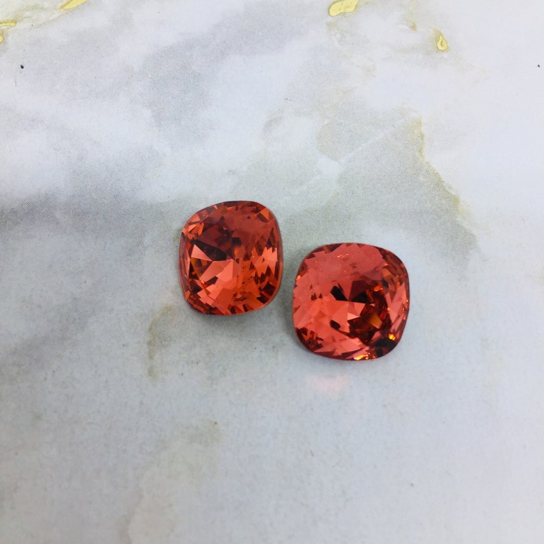 May include: Two orange-red square-shaped rhinestones with a faceted design.