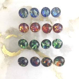 Op de afbeelding: Zestien ronde glazen cabochons gerangschikt in een vier-bij-vier raster. Elke cabochon heeft een glad, gewelfd oppervlak met ingebedde iriserende vlokken in verschillende kleuren, waaronder blauw, rood, groen en donkere tinten met veelkleurige spikkels.
