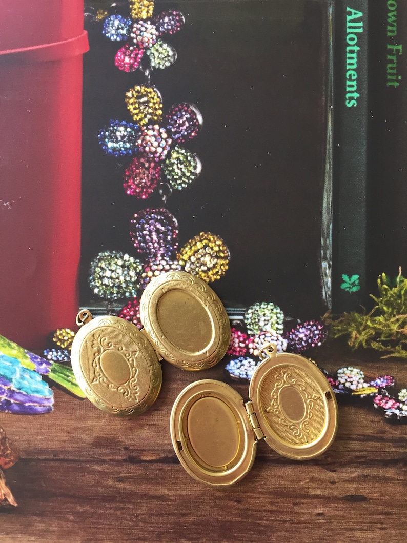 May include: Three gold oval-shaped locket pendants with intricate designs. The lockets are open and show the inside of the locket. The lockets are on a wooden surface.