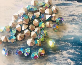 Può includere: Una collezione di strass iridescenti e perline sfaccettate color argento. Gli strass mostrano uno spettro di colori, tra cui blu, verdi e viola. Le perline sono di colore argento chiaro. Gli articoli sono sparsi su uno sfondo blu e bianco.