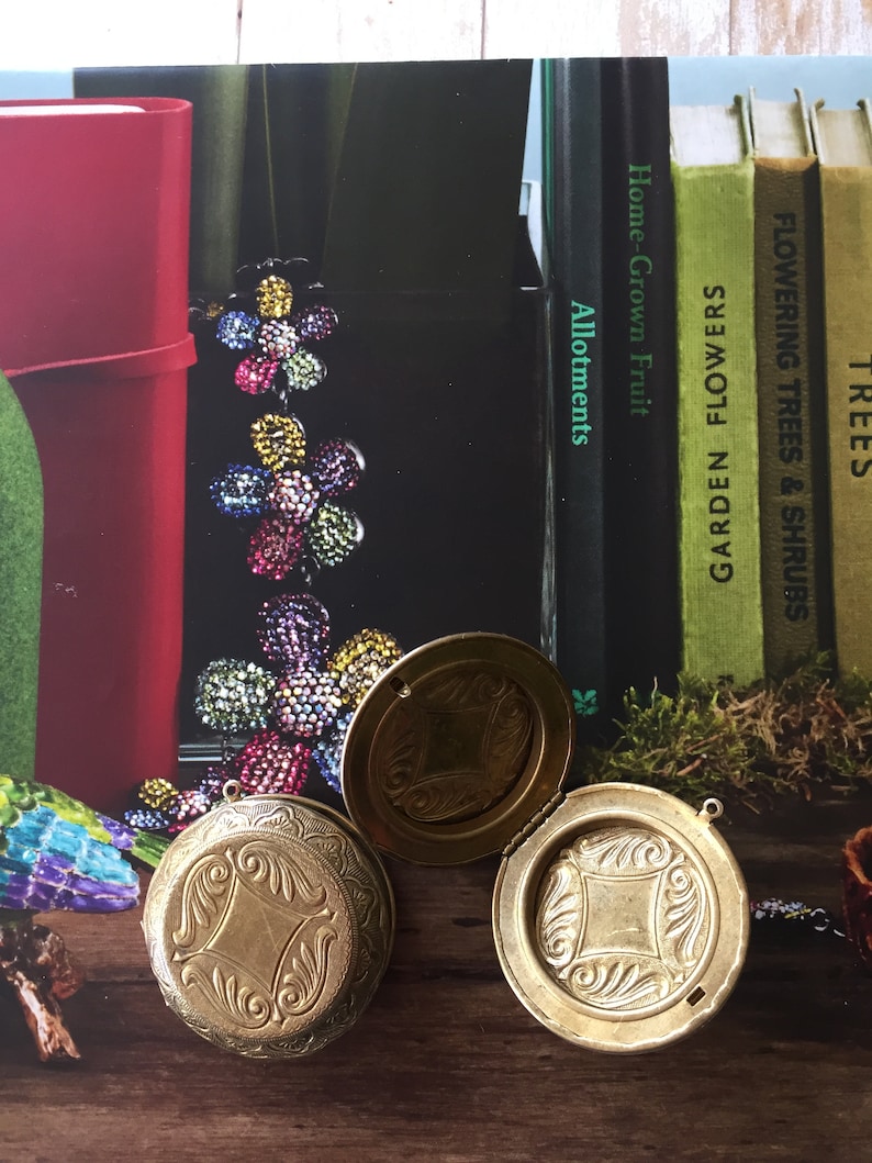 May include: Two gold-coloured, round, hinged pocket watches with ornate designs on the front. The watches are open and lying on a wooden surface.