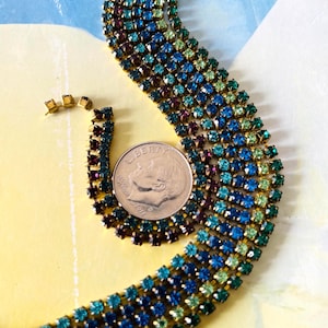 Puede incluir: Un collar vintage de pedrería y pendientes a juego. El collar presenta múltiples filas de pedrería cuadrada de colores en tonos azules, verdes y morados. Los pendientes tienen un diseño similar. Se incluye una moneda como referencia de tamaño.