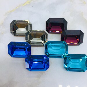 May include: A collection of rectangular-cut gemstones in shades of blue, teal, brown, and deep red. The gems are displayed on a light surface, highlighting their cut and clarity. These gemstones are likely for jewellery making.