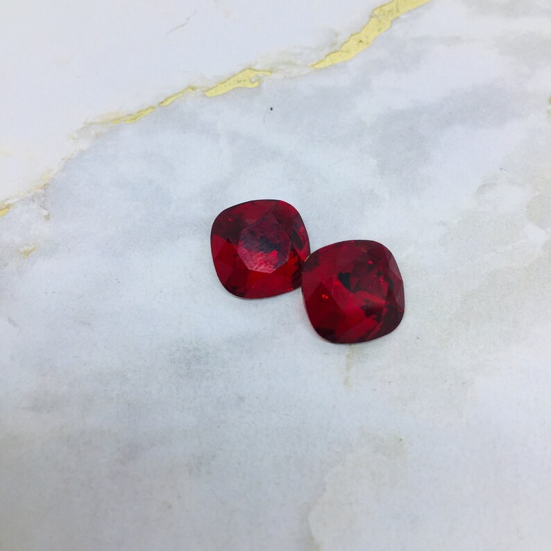 May include: Two red square-shaped rhinestones on a white marble background.