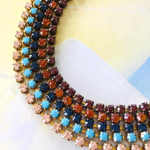 May include: A multi-coloured necklace featuring rows of square gemstones in pink, turquoise, navy, orange, and burgundy. The gemstones are set in a gold-toned metal. A striking piece of jewellery.