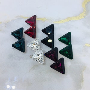May include: Assortment of triangle-shaped rhinestones in various colors, including red, green, black, and clear. The rhinestones are arranged in pairs on a white surface.