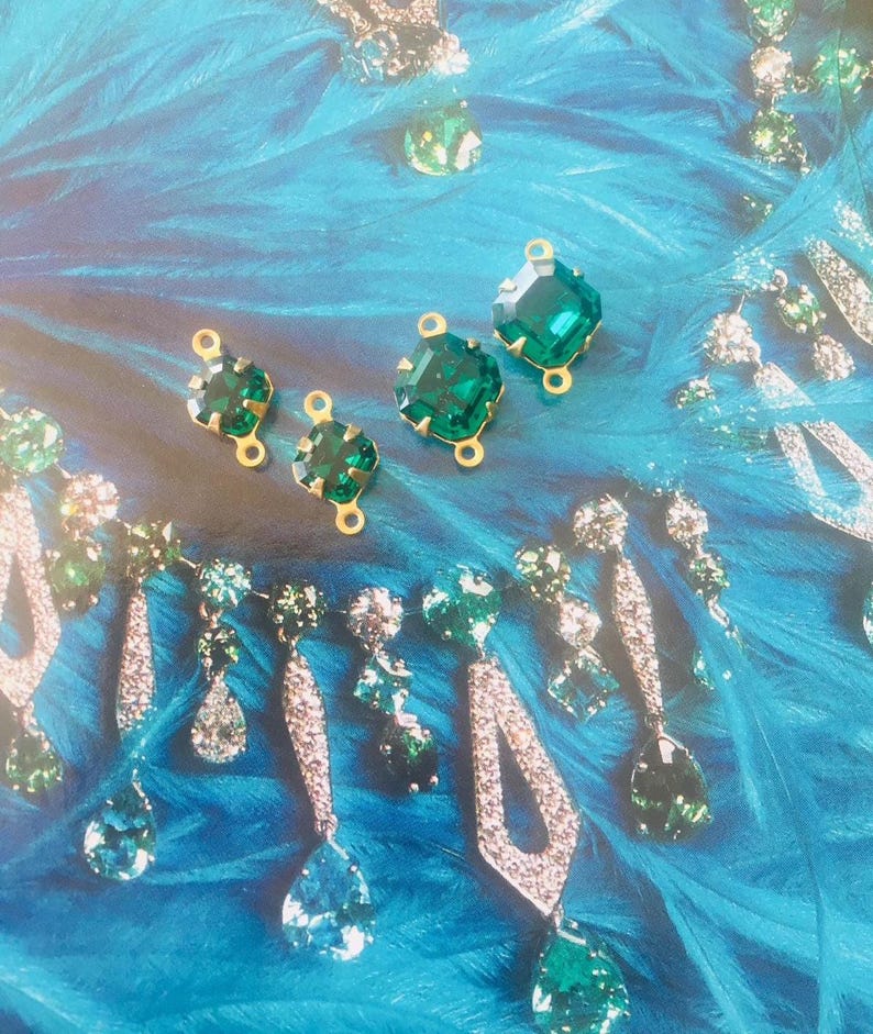 May include: Emerald green gemstone jewelry and components on a blue background. The image features various earrings and loose gemstones with gold settings. The earrings have a mix of clear and green stones. The background has a feathery texture.