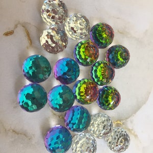 May include: Assortment of iridescent glass beads in various colors, including green, blue, and purple. The beads have a faceted, round shape and are arranged on a white surface.