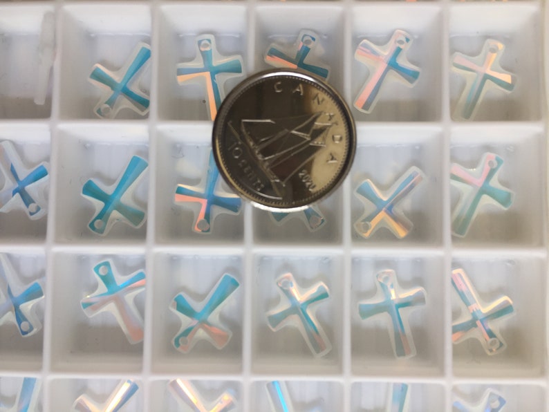 May include: A close-up of a white plastic container filled with iridescent blue and white cross-shaped charms. A Canadian one dollar coin is placed in the center of the container for size reference.