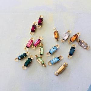 May include: A selection of rectangular crystal charms in various colours, with gold-tone connectors. The crystals include red, pink, green, blue, and orange. These charms are likely for jewellery making or crafting.
