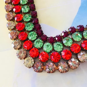 Puede incluir: Collar de varias hebras con filas de piedras preciosas facetadas y coloridas. Las piedras son en tonos rojo, verde, morado y transparente, engastadas en metal dorado. El collar es una pieza llamativa.