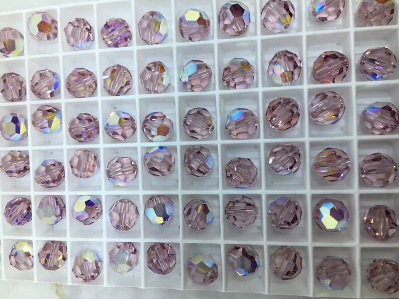 Peut inclure: Vue rapproch&eacute;e d'un plateau de perles de verre de couleur lilas clair. Les perles sont dispos&eacute;es en grille et ont une finition facet&eacute;e et iris&eacute;e.