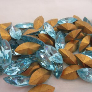 Puede incluir: Un gran grupo de piedras de imitaci&oacute;n de forma marquesa de color azul claro con respaldos dorados. Las piedras de imitaci&oacute;n est&aacute;n esparcidas sobre una superficie blanca.