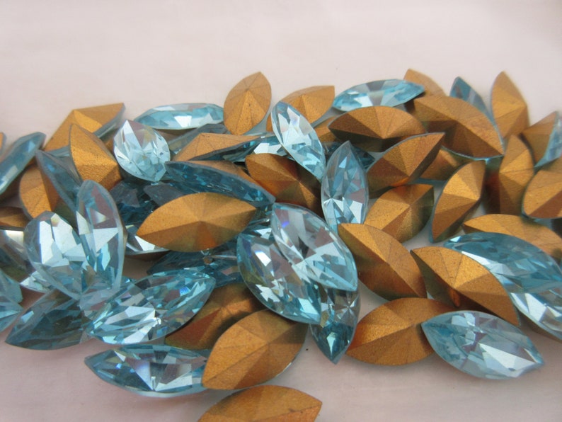 Puede incluir: Un gran grupo de piedras de imitaci&oacute;n de forma marquesa de color azul claro con respaldos dorados. Las piedras de imitaci&oacute;n est&aacute;n esparcidas sobre una superficie blanca.