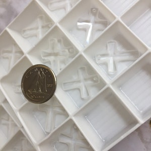 May include: A white plastic tray with multiple compartments filled with clear glass cross charms. A Canadian 10-cent coin is placed in one of the compartments for size reference.
