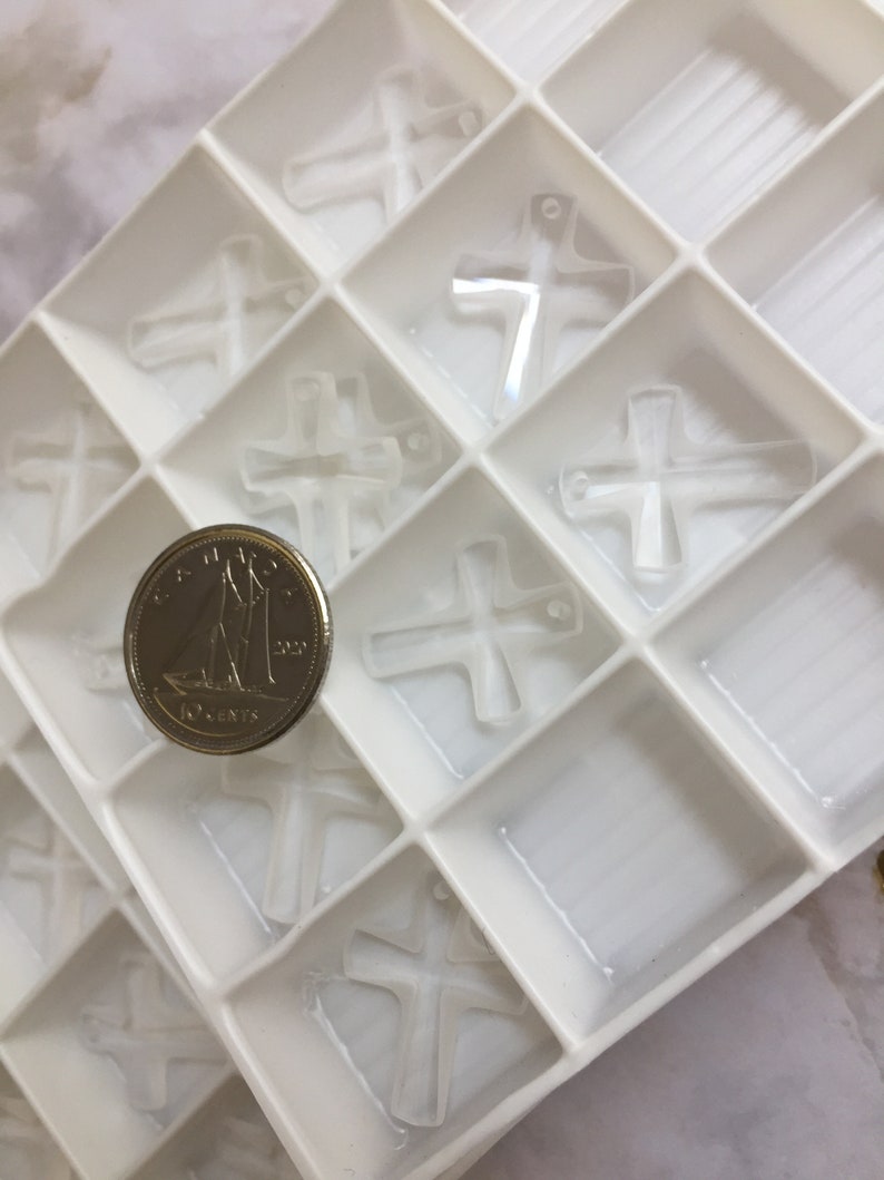 May include: A white plastic tray with multiple compartments filled with clear glass cross charms. A Canadian 10-cent coin is placed in one of the compartments for size reference.