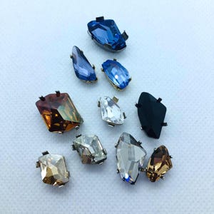 Può includere: Una collezione di abbellimenti di pietre preziose assortite in varie forme e colori, tra cui blu, trasparente, marrone e nero. Ogni pietra ha una montatura in metallo.