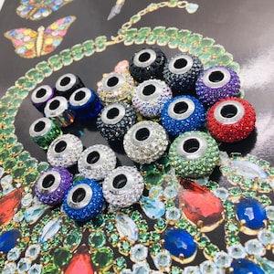 Puede incluir: Una colección de cuentas redondas y coloridas con detalles metálicos plateados. Las cuentas están incrustadas con cristales brillantes en varios colores, incluyendo morado, azul, verde y rojo. Las cuentas se exhiben sobre un fondo decorativo con ilustraciones de joyas y mariposas.