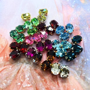May include: A collection of oval-shaped faceted crystals in a variety of vibrant colors including red, green, blue, pink, yellow, and clear. Each crystal is set in a metallic prong setting, creating a cluster of small, reflective, and polished gems.