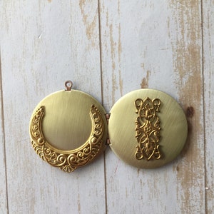 May include: A gold locket with an intricate design. The locket is open, revealing a blank interior. The locket is on a white wooden background.