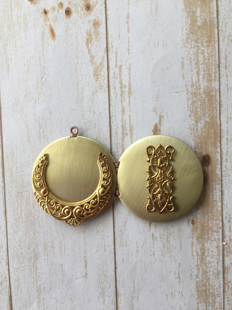 May include: A gold locket with an intricate design. The locket is open, revealing a blank interior. The locket is on a white wooden background.