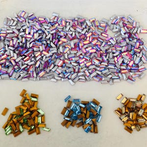 May include: An assortment of rectangular iridescent rhinestones in various colors. The main pile is silver with rainbow hues. Smaller piles of orange, blue, and gold rhinestones are also present, suitable for crafts and embellishments.