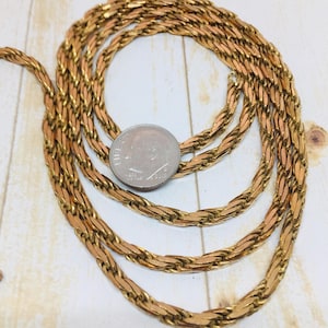 May include: A gold-toned chain necklace with a woven design, coiled on a light wood surface. A US dime is placed on the chain for size reference. The necklace has a warm, metallic color.