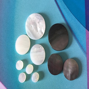 May include: Assortment of round and oval shaped mother of pearl and black shell buttons. The buttons are various sizes and are arranged on a blue and purple background.